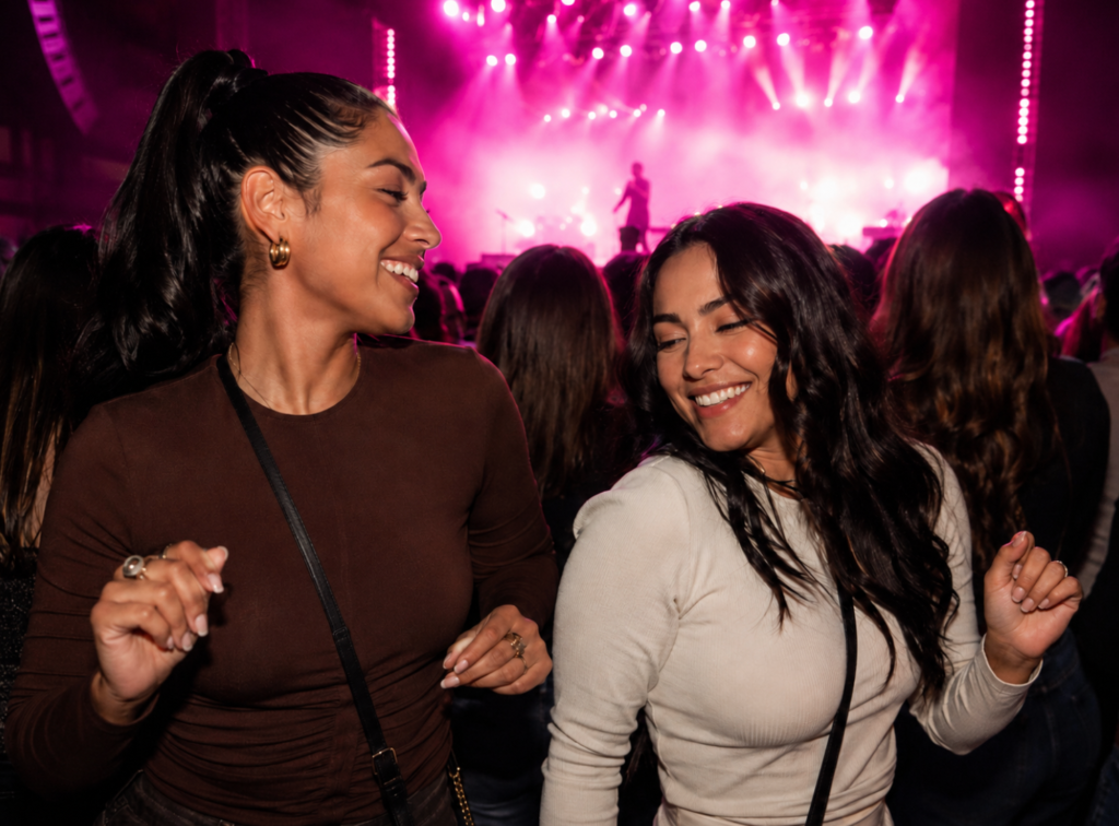 Dos amigas bailando y sonriendo en un concierto con luces rosadas y una cantante en silueta sobre el escenario.
