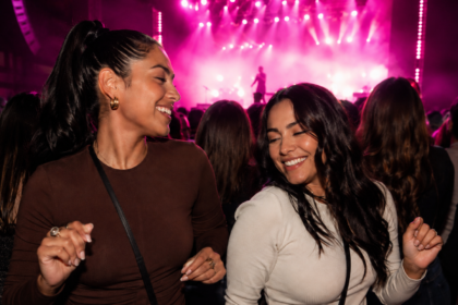 Dos amigas bailando y sonriendo en un concierto con luces rosadas y una cantante en silueta sobre el escenario.